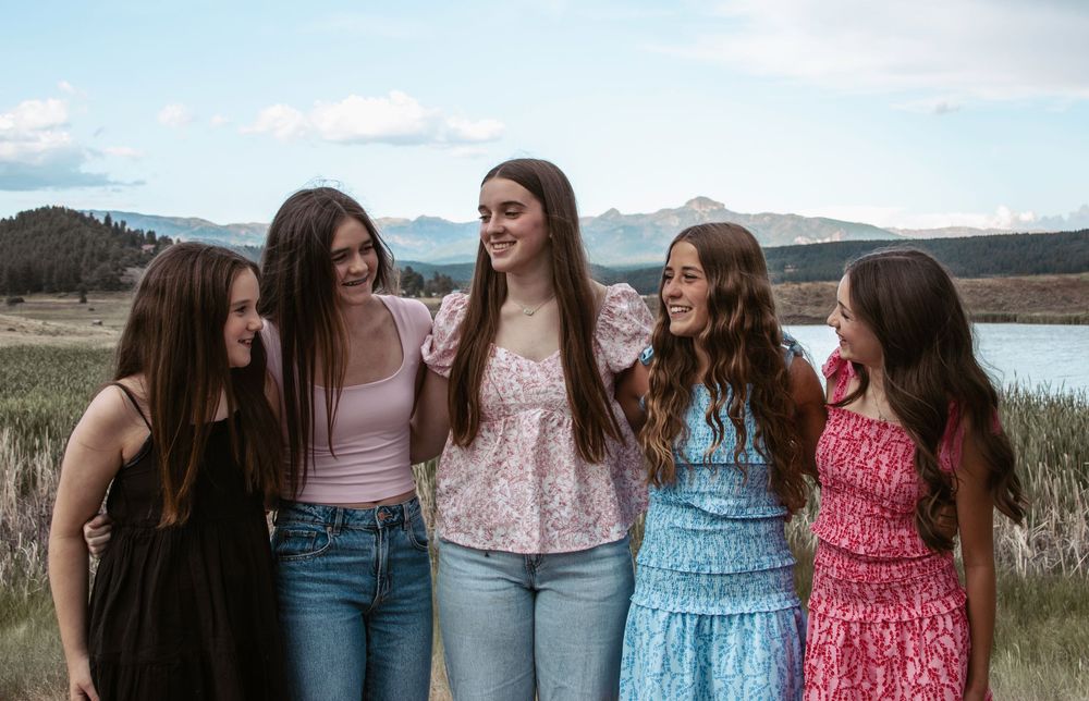 A group of friends in casual dresses and jeans stand together by a scenic lake with mountains in the background.