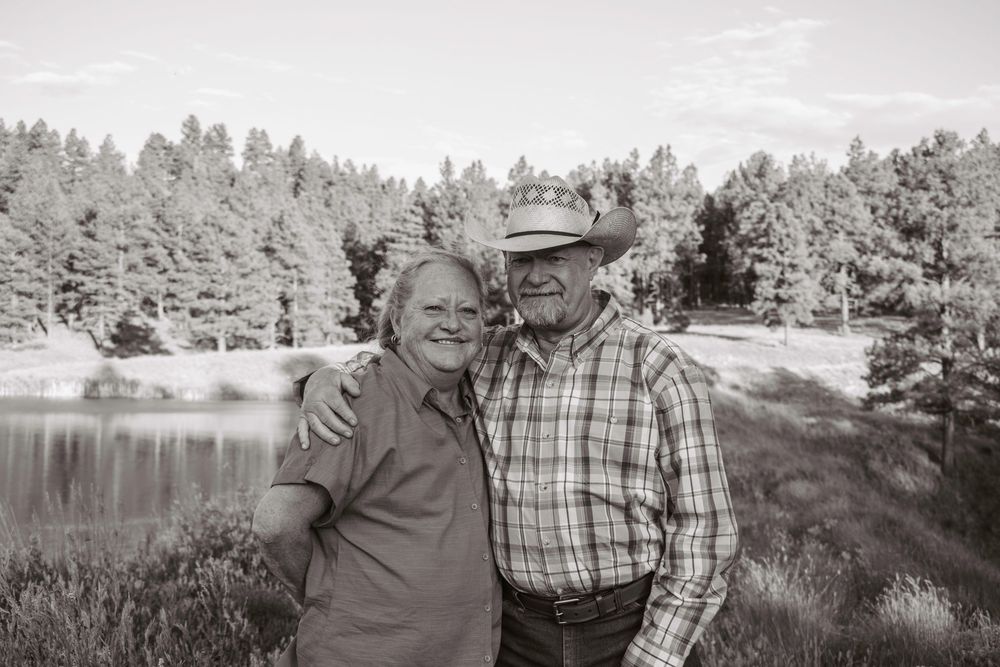 A couple stands together outdoors by a serene lake surrounded by pine trees in black and white.
