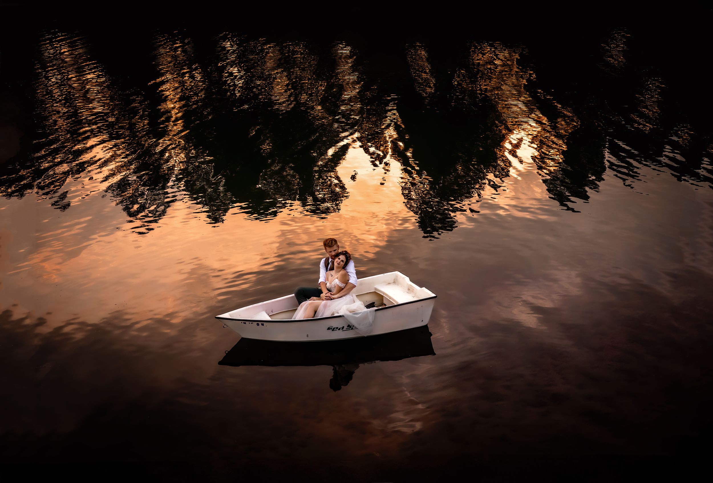 Bride and groom sit in a small white boat on reflective water at sunset with silhouetted trees in the background.