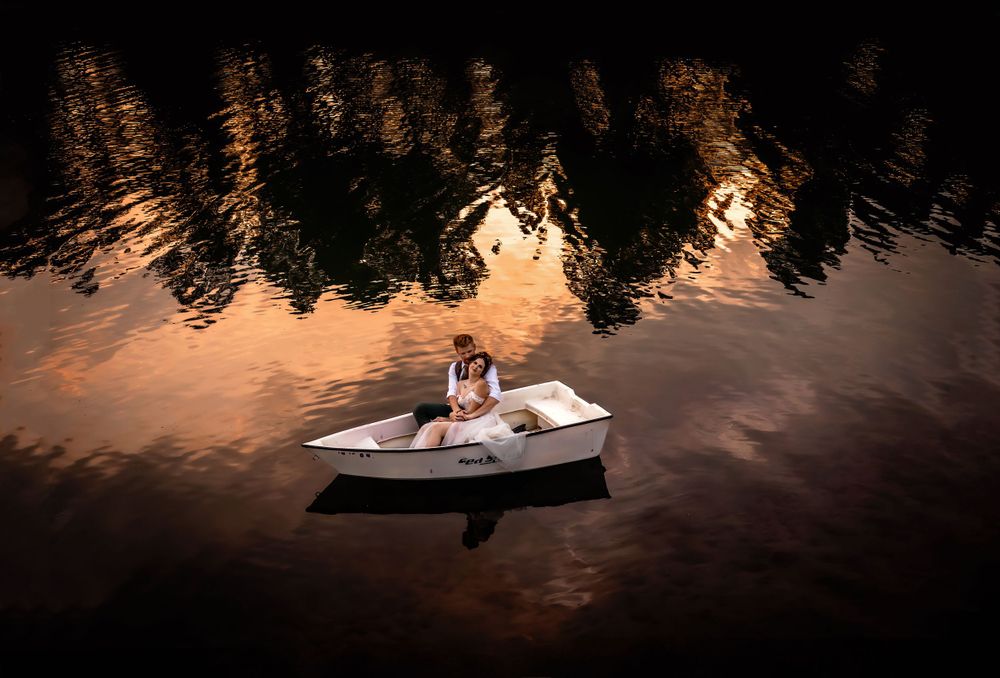 Bride and groom sit in a small white boat on reflective water at sunset with silhouetted trees in the background.