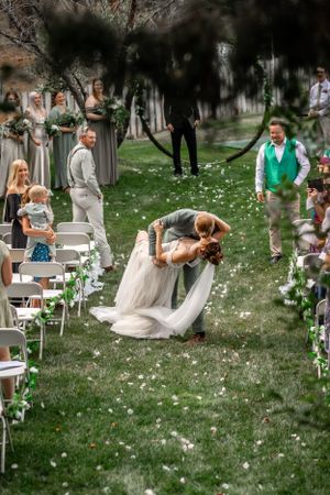 Wedding couple shares romantic dip kiss during outdoor ceremony while guests watch and flower petals float through the air.