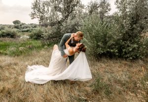 A romantic couple in formal attire share a dip pose in a rustic outdoor field with green trees in the background.