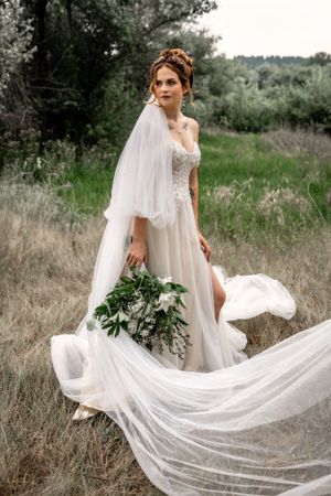 Elegant bride in flowing white gown with dramatic veil holds greenery bouquet while standing in rustic field setting.