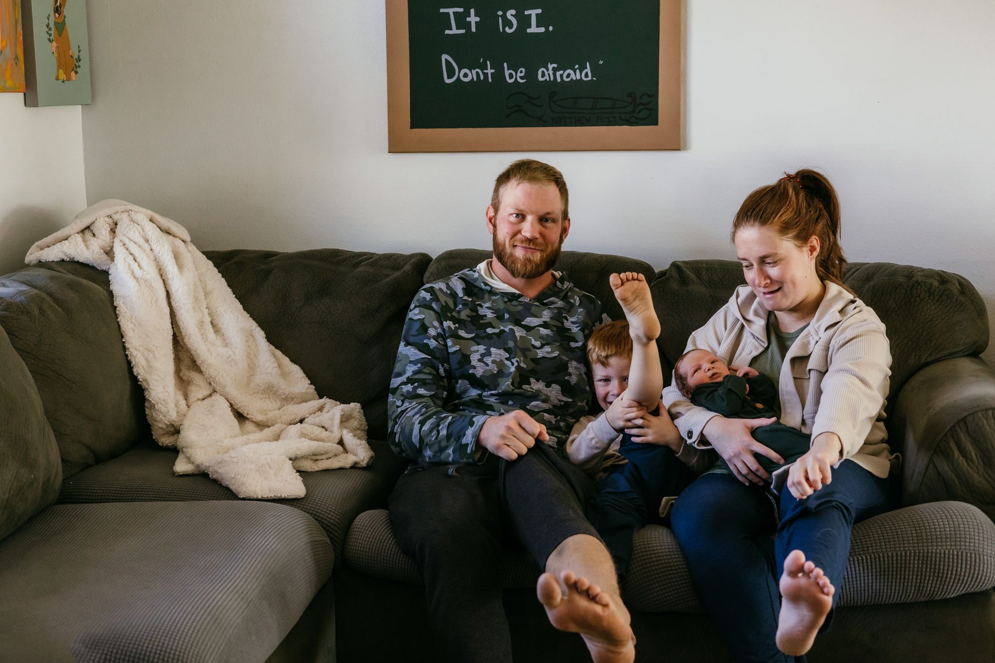 Family sits relaxed on a gray sectional sofa in a living room with a chalkboard on the wall in the background.