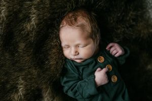 Sleeping newborn wearing dark green sweater rests peacefully on soft brown fur blanket.
