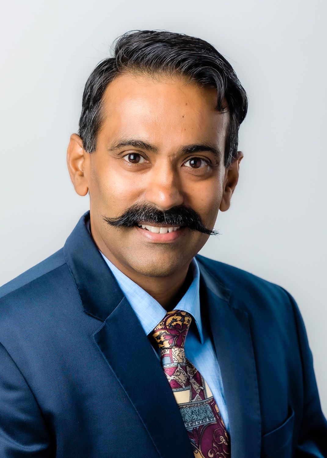 Professional male Linkedin profile with moustache wearing a navy suit and patterned tie posing confidently against a light background