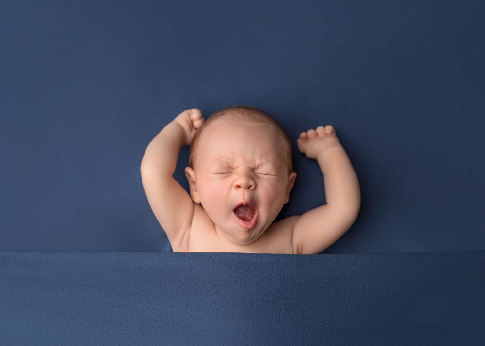 Newborn yawning while lying on dark blue backdrop.