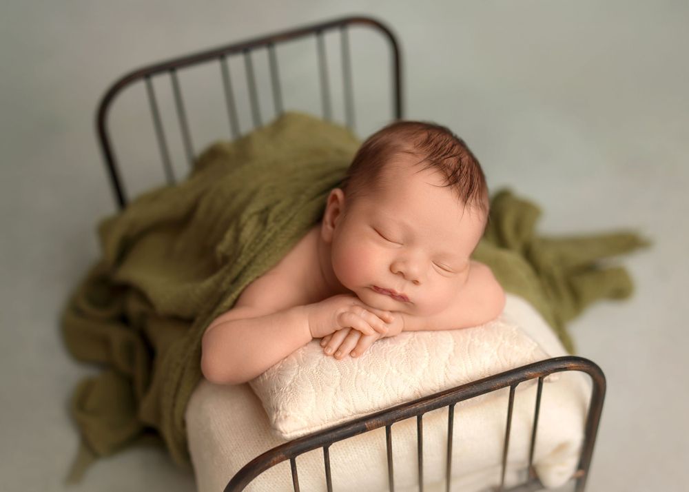 Sleeping newborn posed on antique metal bed with olive green textile.