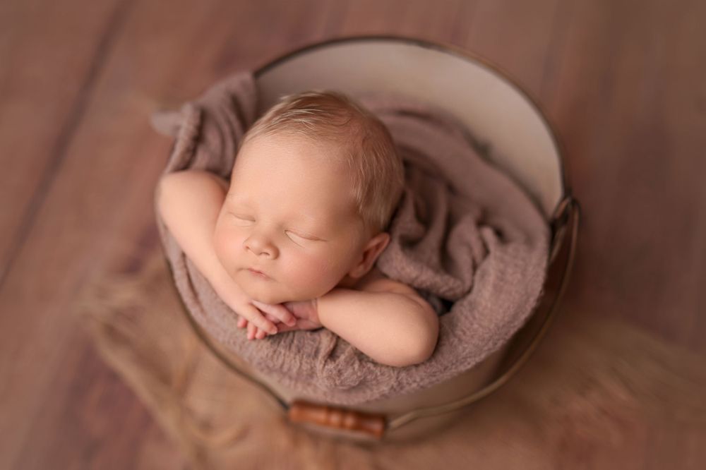 Sleeping newborn curled up in round brown container with neutral-toned wrapping.