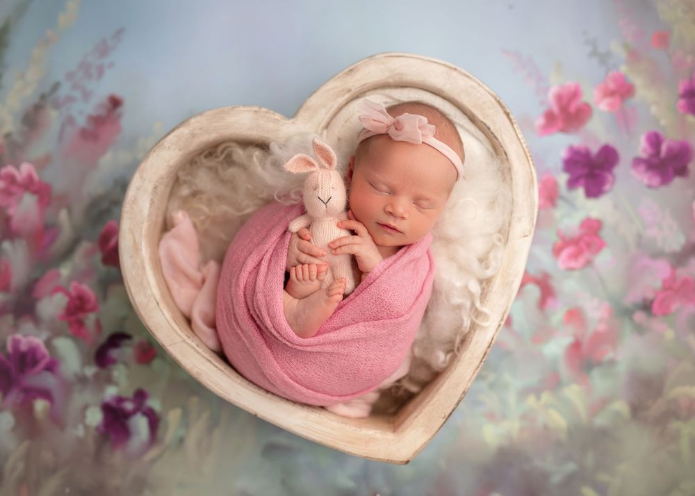A sleeping newborn wrapped in pink swaddle rests in a heart-shaped bowl against a floral backdrop.
