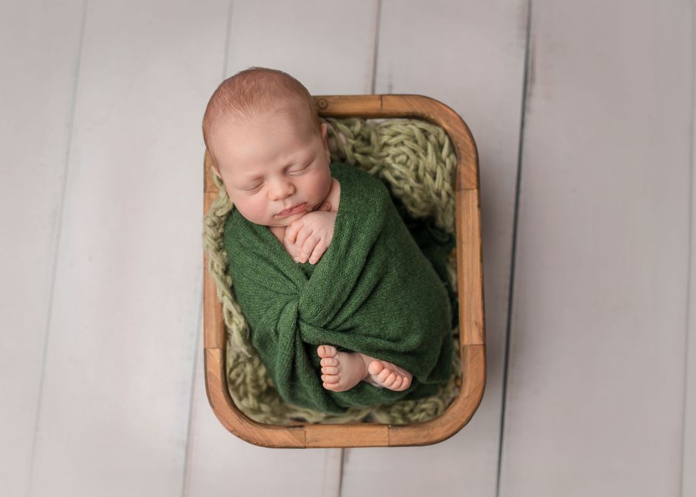 A sleeping newborn wrapped in green blanket lying in wooden basket on grey backdrop.