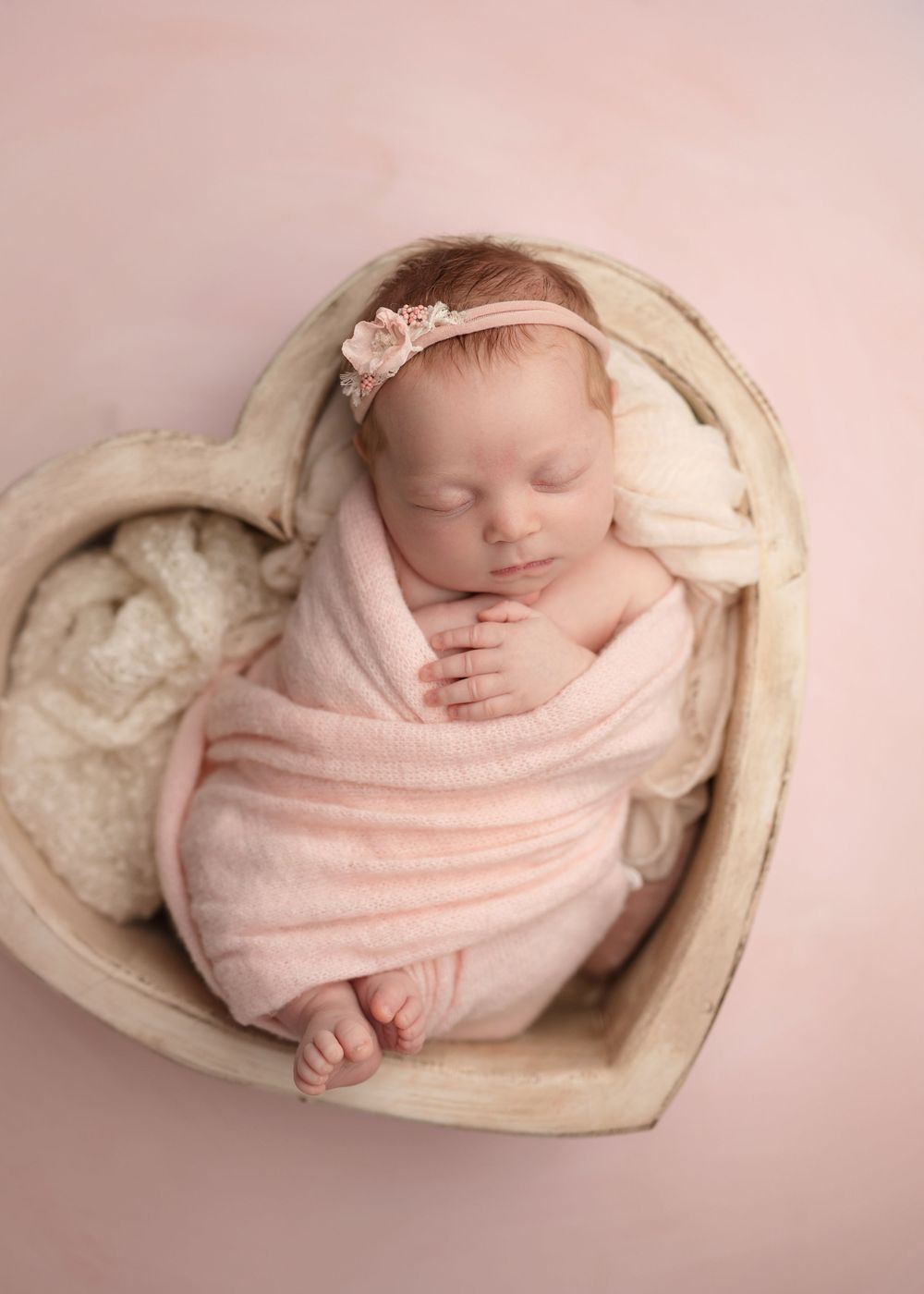 Newborn baby sleeping wrapped in pink swaddle blanket nestled in cream-colored heart-shaped basket.