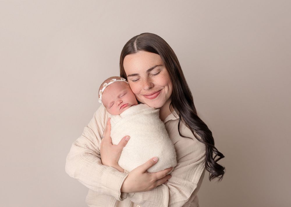 A tender moment between parent and sleeping newborn baby, both wearing soft neutral colors against beige backdrop.