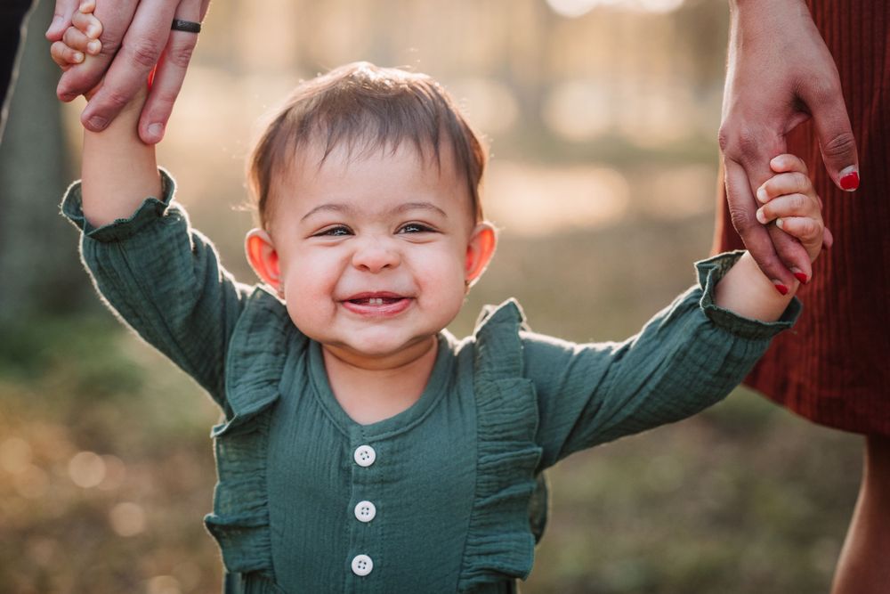 Baby in green outfit reaching up while learning to walk outdoors.