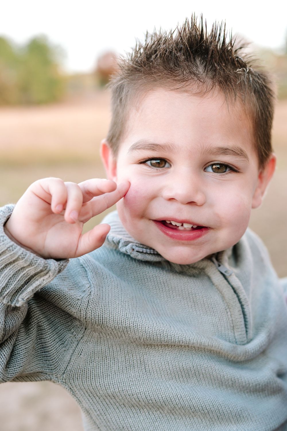 Child wearing gray sweater points to cheek while smiling outdoors.