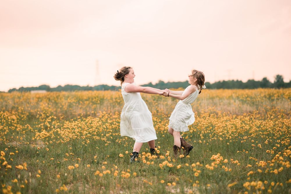 Two children in white dresses dancing together in a field of yellow wildflowers at sunset.