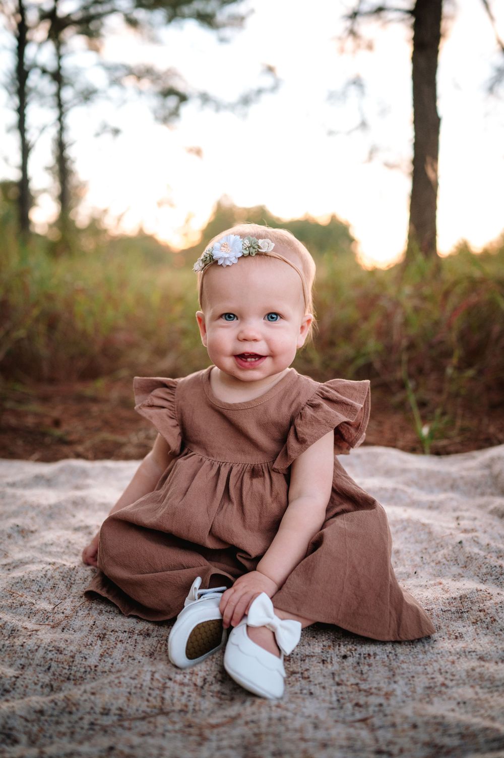 Baby wearing brown dress and white shoes sitting on sandy ground during golden hour photo session.