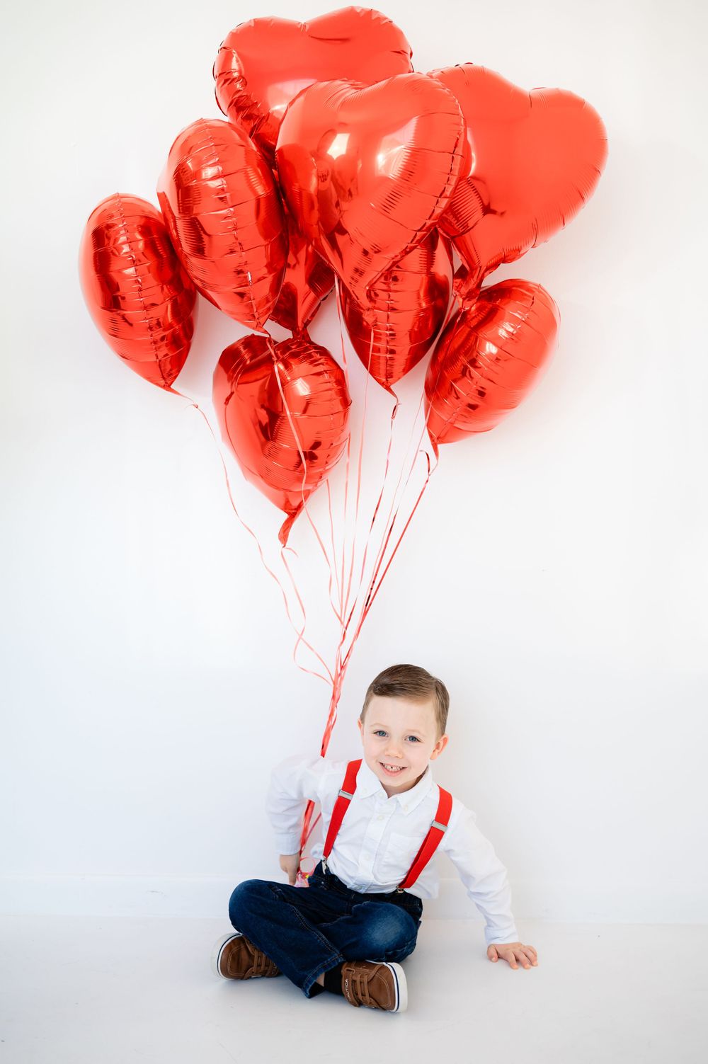 Child sitting cross-legged holding red heart-shaped balloons against white background.