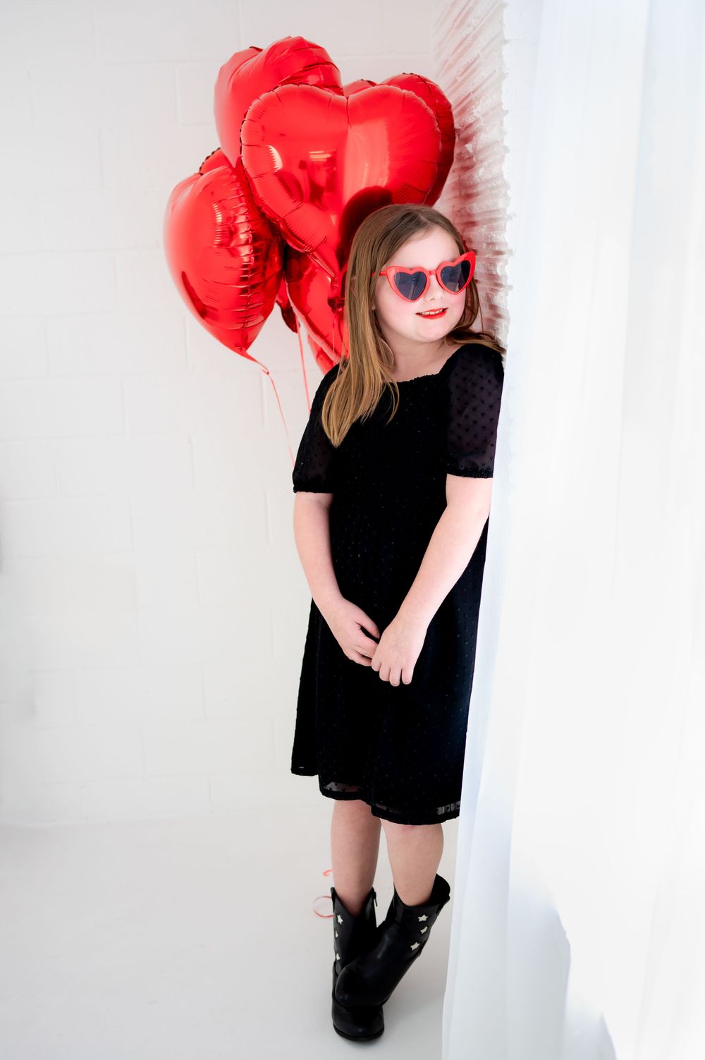 Child in black dress and heart sunglasses poses with red heart balloons against white background.