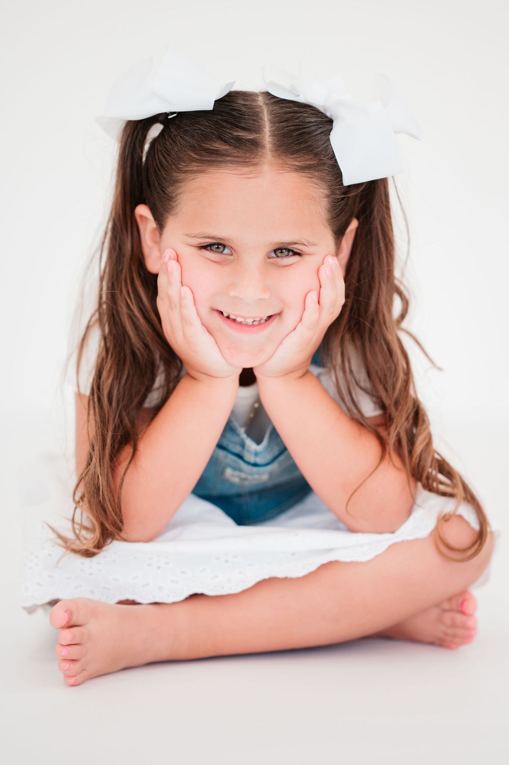 Child with pigtails resting head in hands while lying on white background.