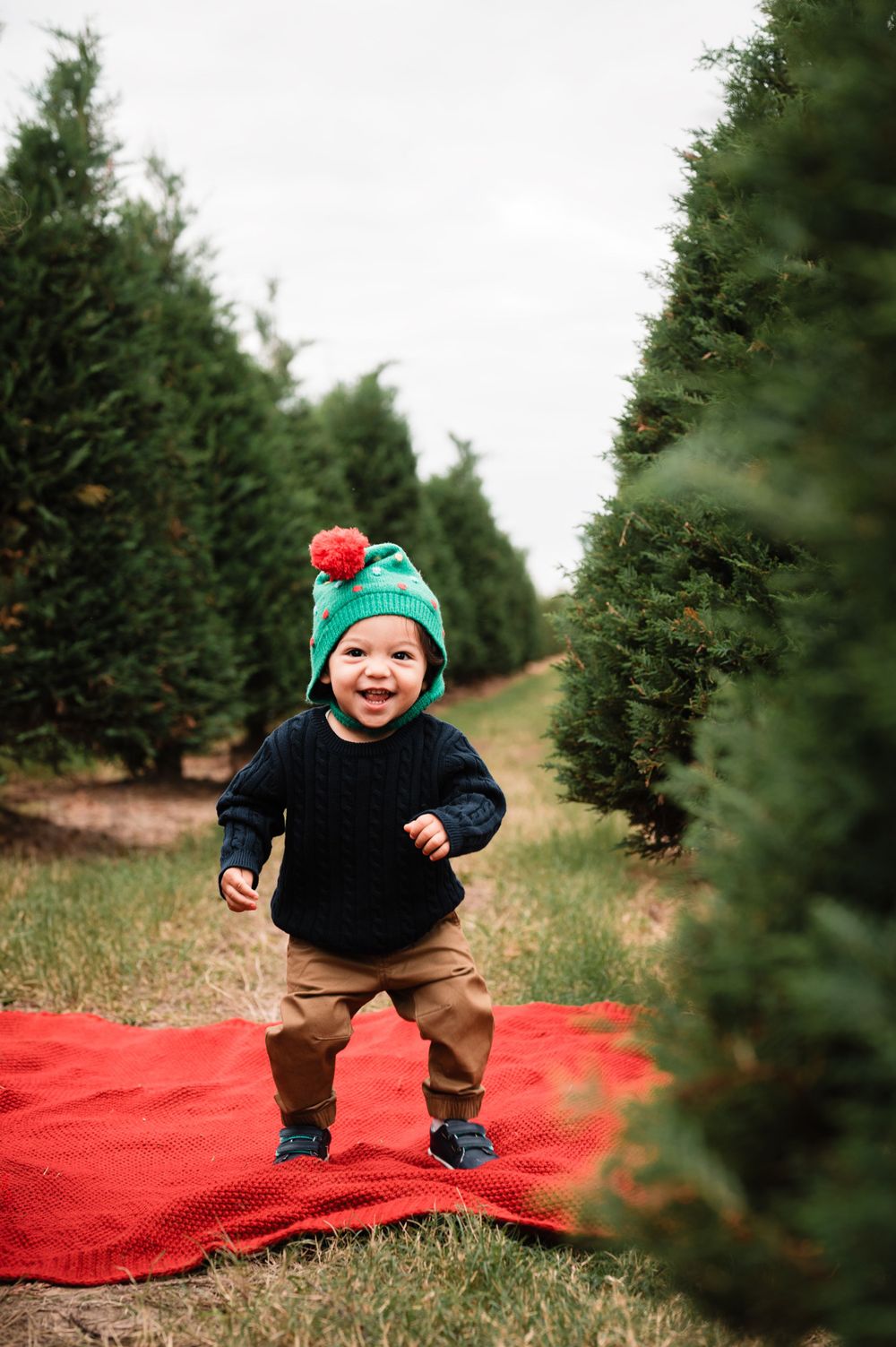 Child jumping on red blanket between Christmas trees during outdoor holiday photo shoot.