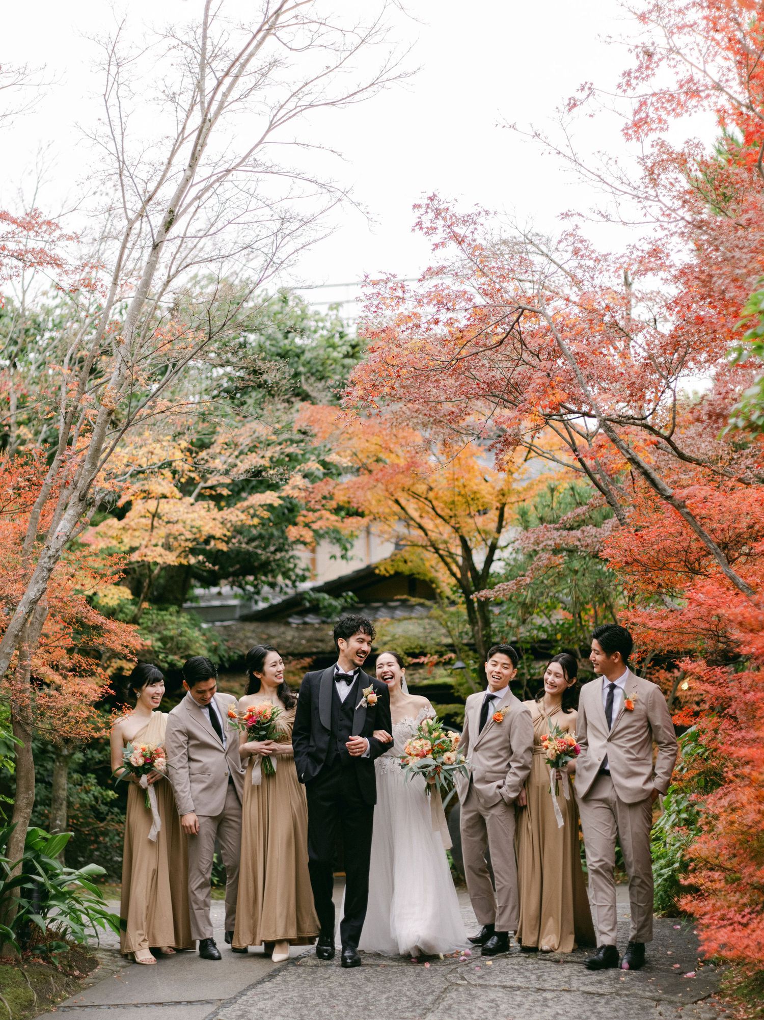 Wedding party in autumn garden with bride, groom, and attendants in gold dresses and tan suits amid vibrant fall foliage.