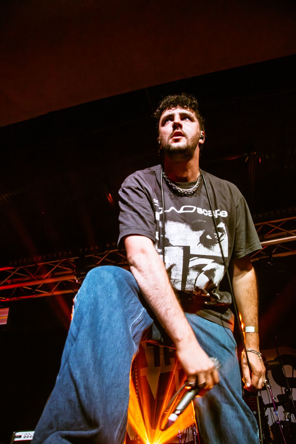Performer in blue jeans and t-shirt captured during dramatic low-angle concert shot with red lighting.