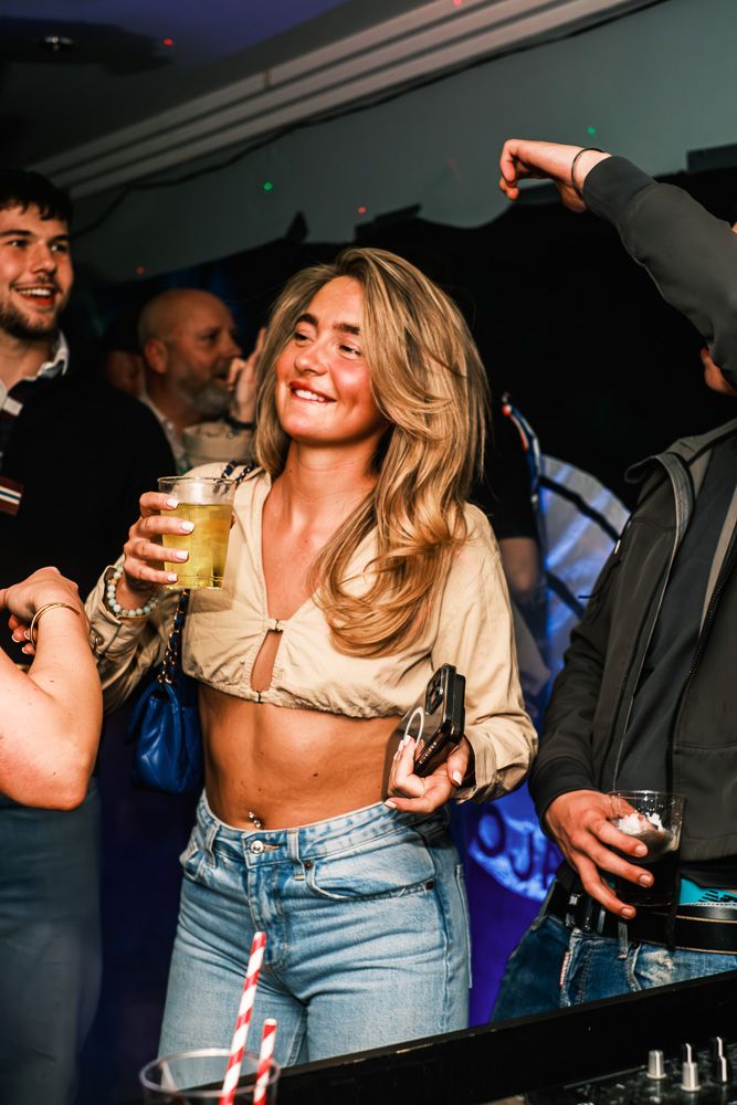 Person in gold top and jeans dances with drinks at a crowded nightclub party, creating an energetic party atmosphere.