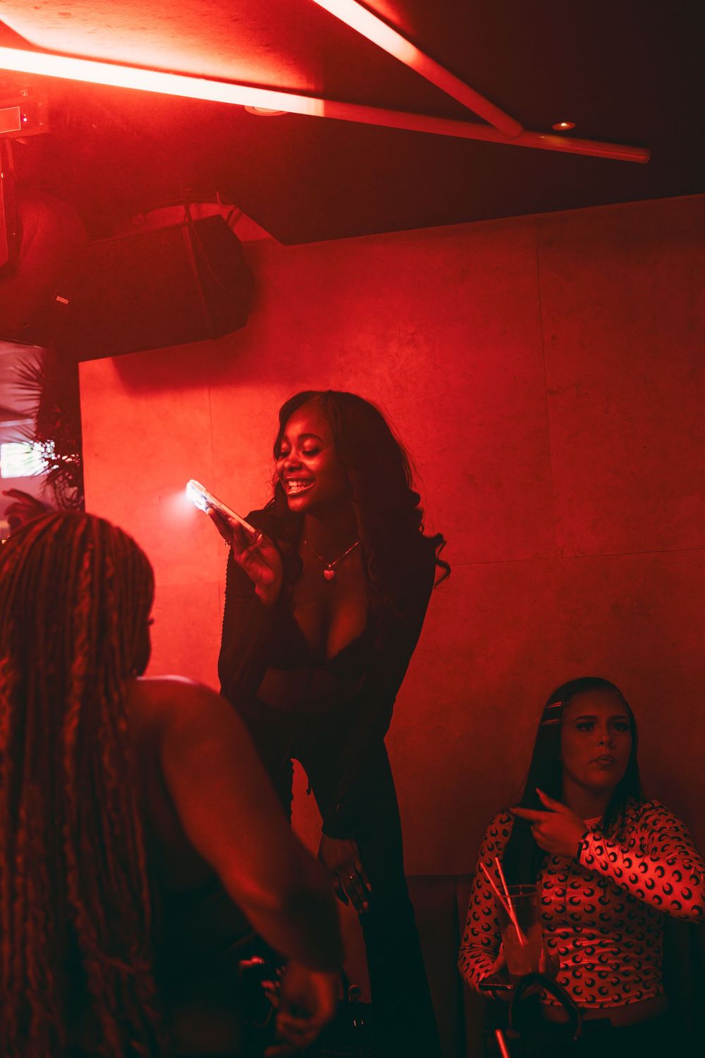 A moody nightclub scene bathed in red lighting shows silhouettes of people dancing and socializing in a hazy atmosphere.