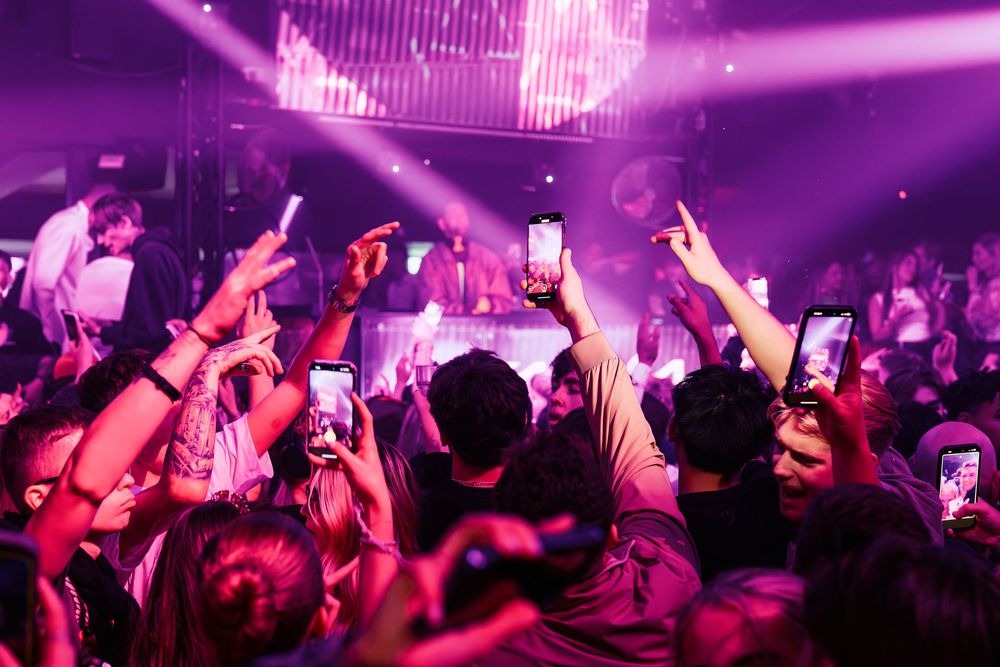 Energetic crowd with raised hands dancing at a nightclub under vibrant purple lighting and disco effects.