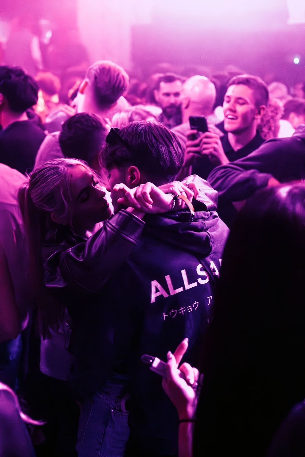 A crowd of concert-goers dance and celebrate under pink and purple lighting at a nightclub event.