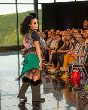A model walks the runway wearing a green skirt and black top at a fashion show with audience members seated along the sides.