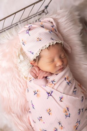 A sleeping newborn swaddled in a pink floral patterned blanket rests peacefully on soft white fur bedding.