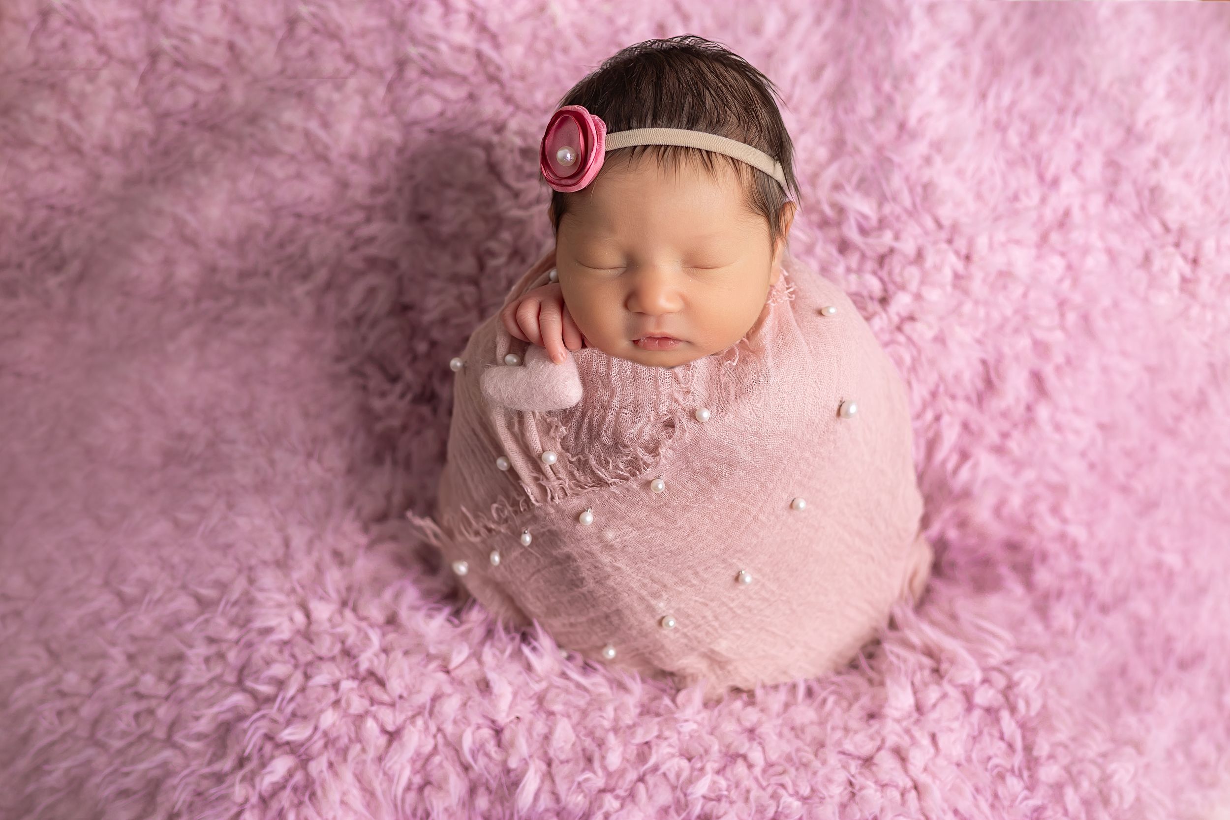 Newborn baby sleeping peacefully on soft pink fluffy blanket with delicate headband accessory.
