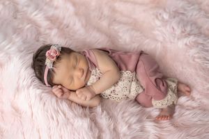 A sleeping newborn baby wearing a lace headband lies on soft pink fur fabric.