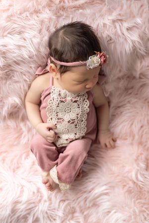 Newborn baby wearing pink outfit with lace detail lies on soft pink fur blanket in professional photo shoot setting.