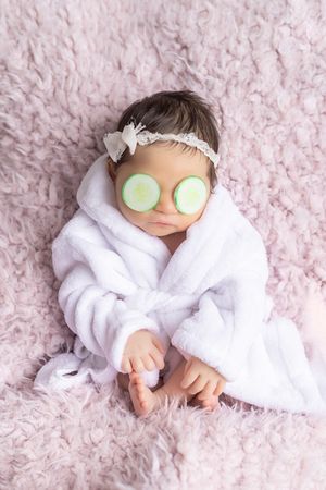 A baby in a white robe relaxes with cucumber slices on eyes during a spa-themed newborn photo session on pink blanket.