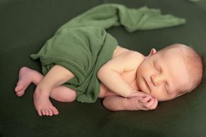 Sleeping newborn baby in green wrap lying on dark green backdrop during professional photo session.
