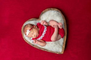 Sleeping newborn in red outfit posed on heart-shaped cushion against red background.