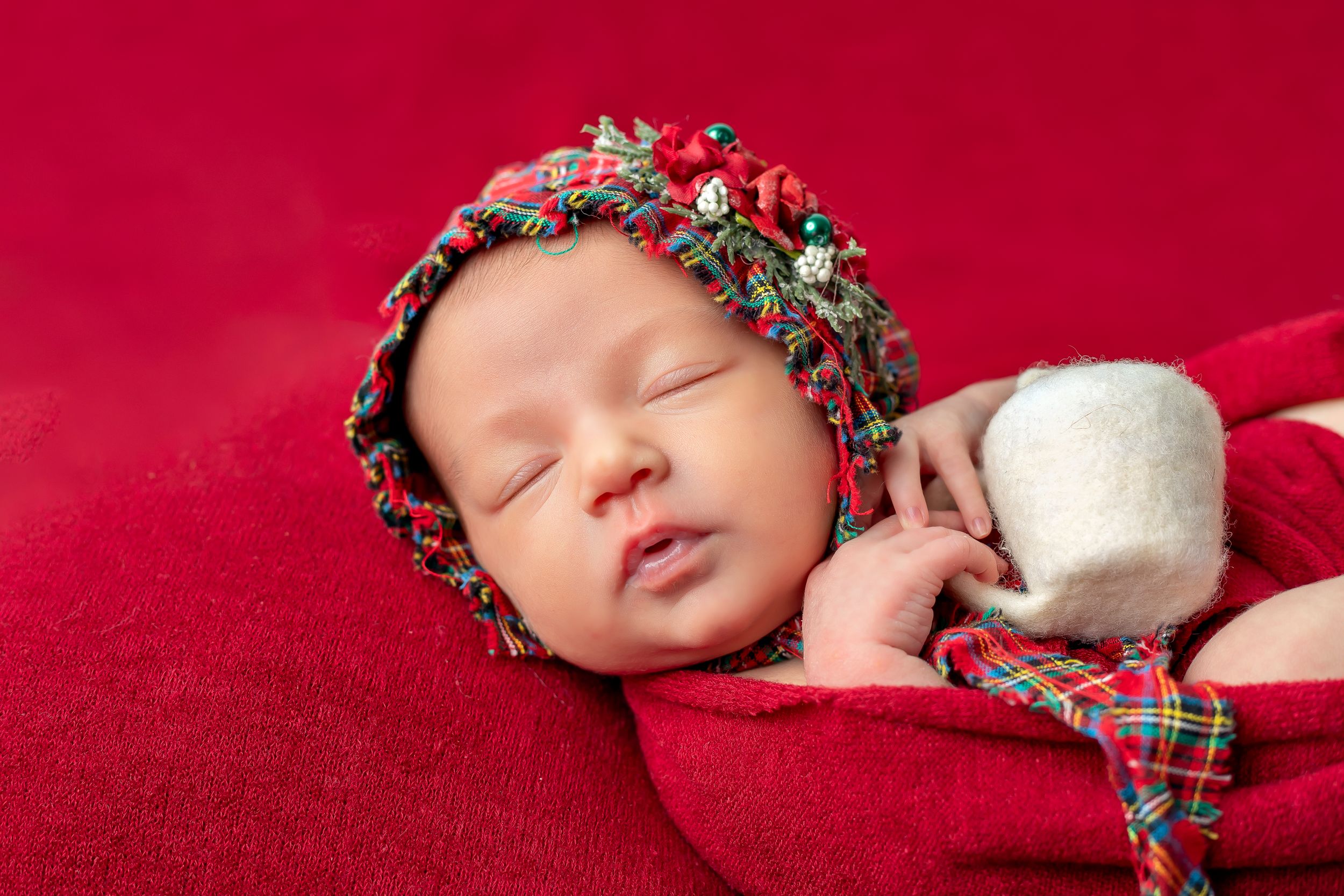 Sleeping newborn wearing plaid bonnet on red velvet backdrop.
