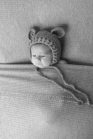 Newborn wearing gray knitted bear bonnet lying on textured gray fabric in black and white.