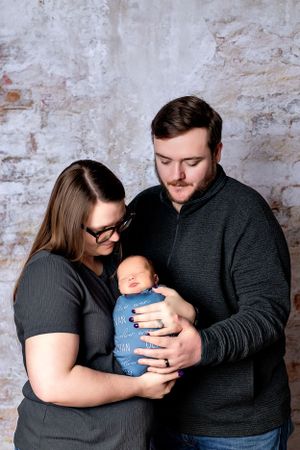 Parents in dark clothing tenderly hold their sleeping newborn baby swaddled in blue against a rustic brick wall.