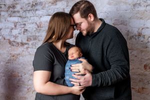 Parents in dark clothing holding their newborn baby wearing blue against a rustic brick wall background.