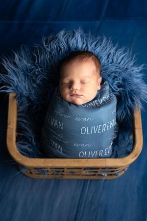 A sleeping newborn wrapped in a gray personalized name blanket rests in a wooden basket with blue fur padding.