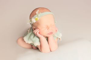 Sleeping newborn in mint green outfit and floral headband posed on beige backdrop.