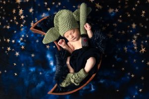 A sleeping baby wearing a knitted green hat and bootees lies in a wooden moon prop against a starry blue background.