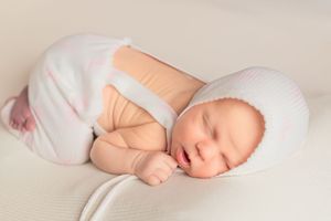 A sleeping newborn baby in white bonnet and peach outfit curled up on cream-colored blanket.