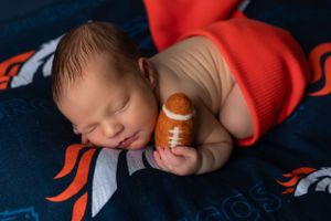 Sleeping baby in orange outfit holds tiny football while resting on blue and orange sports team blanket.