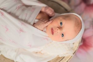 A baby with bright blue eyes wearing a white bonnet lies on pink bedding.
