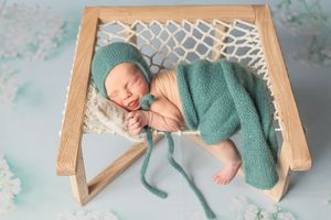 Sleeping newborn wrapped in teal knit blanket rests peacefully in wooden hammock prop during photo session.